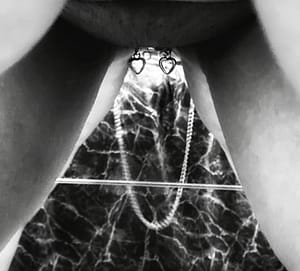 Black and white photo taken in the mirror, perspective is between my tits and thighs. The heart charms on my clit clip dangle above the reflection of the chain between my nipple clamps, making a happy face with heart-shaped eyes.