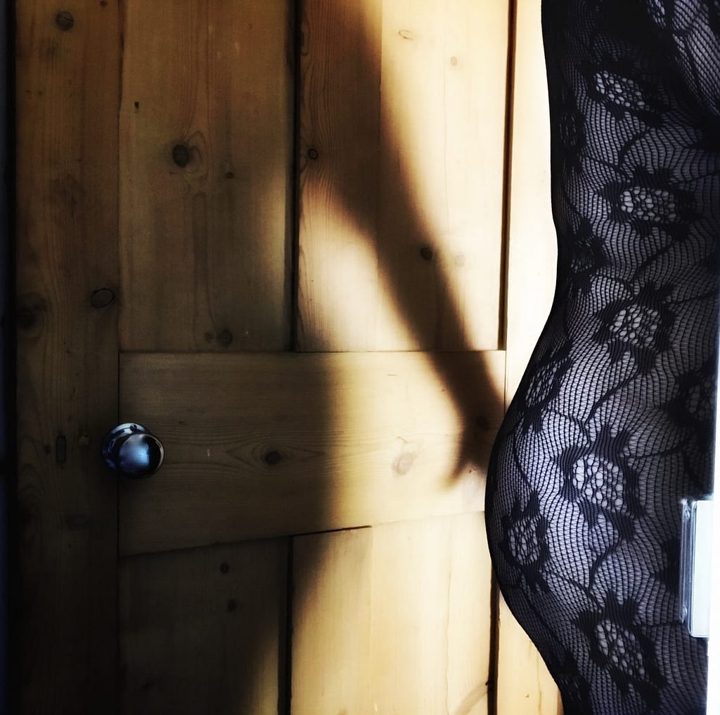 Image shows my back and bum at the edge of the door frame, with my shadow on the wooden door behind me, arm outstretched  so that my hand is on my bum. I’m wearing a flowery lace bodystocking 